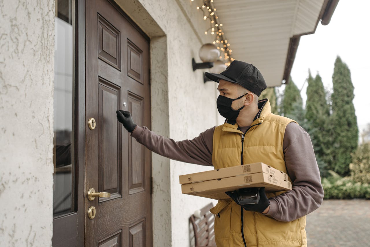 A delivery man wearing a mask knocks on a door holding pizzas, symbolizing contactless delivery during the pandemic.